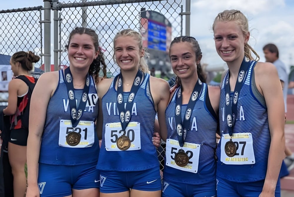 Vier Schülerinnen der Cathedral Preparatory School stehen in Sportuniformen neben dem eingezäunten Leichtathletikstadion.