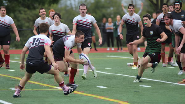 Schüler der Catholic University School spielen Rugby auf einem Rasenfeld mit Zuschauern im Hintergrund.