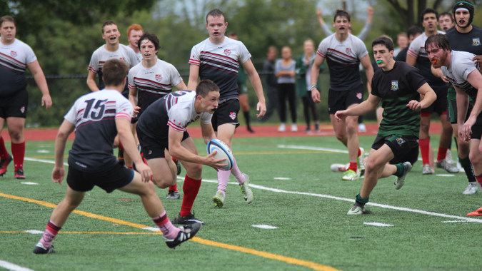 Schüler der Catholic University School spielen ein Rugbyspiel auf einem Spielfeld mit Bäumen und Gebäuden im Hintergrund.