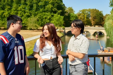 Drei Schüler der CATS Cambridge stehen zusammen und unterhalten sich in einem parkähnlichen Bereich mit Bäumen und Wasser.