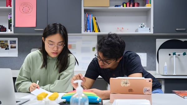 Zwei Personen arbeiten gemeinsam an einem Projekt in einem Raum mit Regalen voller Bücher und Büromaterialien auf dem Campus der CATS Cambridge.