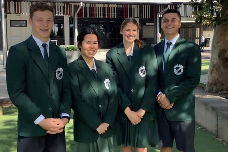 Vier Personen in grünen akademischen Gewändern stehen vor einem Gebäude auf dem Campus der Cavendish Road State High School.