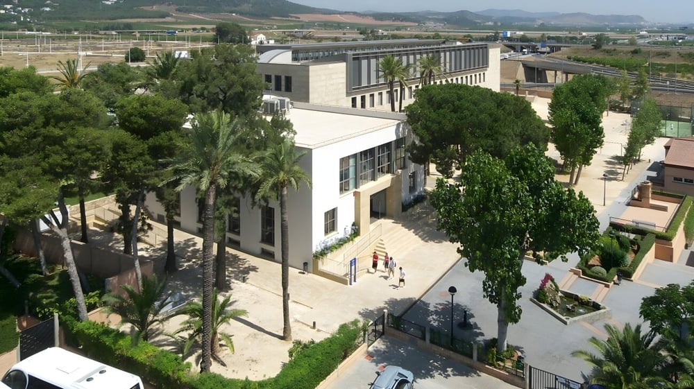 Das moderne mehrstöckige Gebäude des Caxton College ist von grüner Vegetation und Palmen umgeben, mit Menschen auf den Wegen davor.