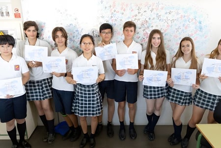 Schüler des Caxton College stehen im Klassenraum zusammen und halten Zertifikate vor einer bunten Wandtafel.