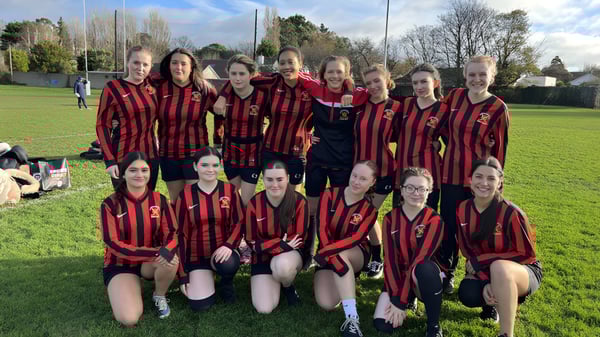Eine Gruppe junger Fußballspielerinnen der CBS Westland Row posiert auf einem Rasenplatz mit Bäumen und Gebäuden im Hintergrund.