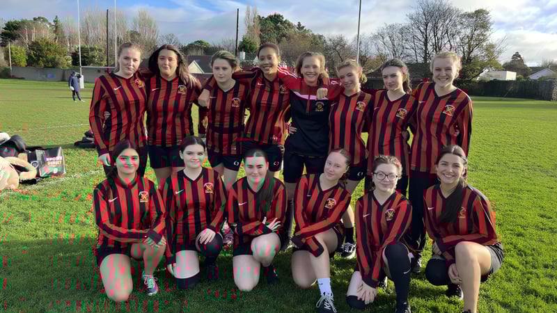 Eine Gruppe junger Fußballspielerinnen der CBS Westland Row posiert auf einem Rasenplatz mit Bäumen und Gebäuden im Hintergrund.