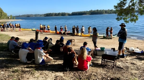 Eine Gruppe von Schülerinnen und Schülern der Centenary Heights State High School versammelt sich am Seeufer inmitten von Bäumen.