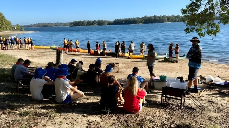 Eine Gruppe von Schülerinnen und Schülern der Centenary Heights State High School versammelt sich am Seeufer inmitten von Bäumen.