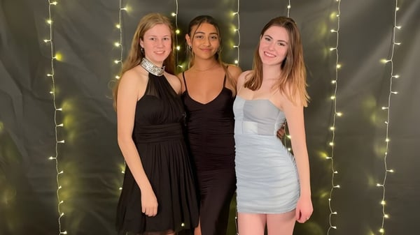 Drei Schülerinnen in formeller Kleidung posieren vor funkelndem Lichterhintergrund auf dem Campus der Centennial Collegiate.