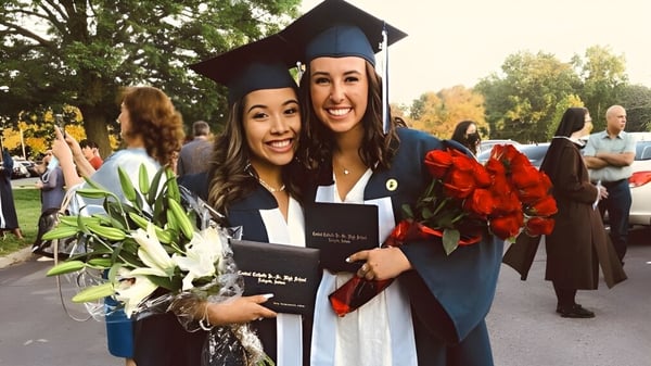 Zwei Schülerinnen der Central Catholic Jr.-Sr. High School in Abschlusskleidung halten Blumen und lächeln vor dem Hintergrund weiterer Absolventen.