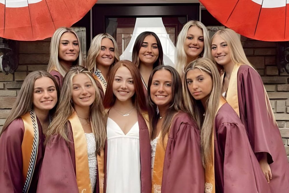 Eine Gruppe von Absolventinnen der Central Regional High School steht in Burgundy-Abschlussroben vor einem Backsteingebäude mit roten und weißen Dekorationen.