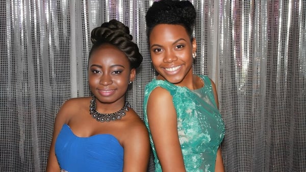 Zwei Frauen mit bunten Frisuren und Kleidern posieren vor einem metallischen Vorhang auf dem Campus der Central Technical School.