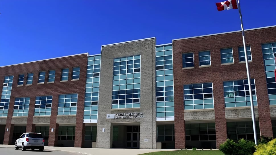 Das mehrstöckige Backsteingebäude der Centre Wellington District High School mit großen Fenstern und einer kanadischen Flagge auf dem Dach im Sonnenschein.
