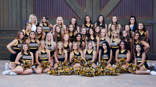 Eine Gruppe Cheerleader mit Pompons posiert gemeinsam vor einem Holzgebäude auf dem Gelände des Chaffey Joint Union High School District.