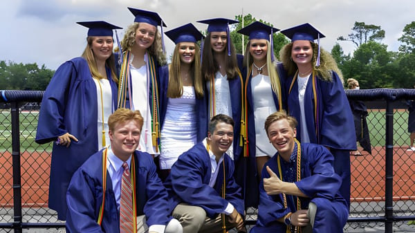Eine Gruppe von Absolventinnen und Absolventen der Chatham Kent Secondary School steht vor einem umzäunten Sportplatz.