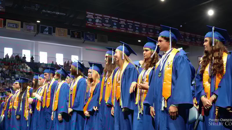 Eine Gruppe von Absolventen in blauen und goldenen Talaren steht auf der Bühne der Chelsea High School während der Abschlussfeier.