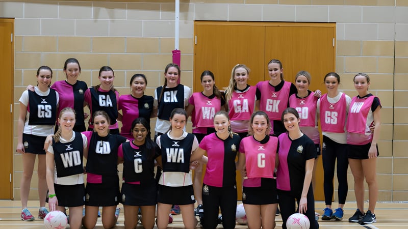 Eine Gruppe junger Netballspielerinnen des Cheltenham College posiert in der Sporthalle mit Holzfußboden und -wänden.