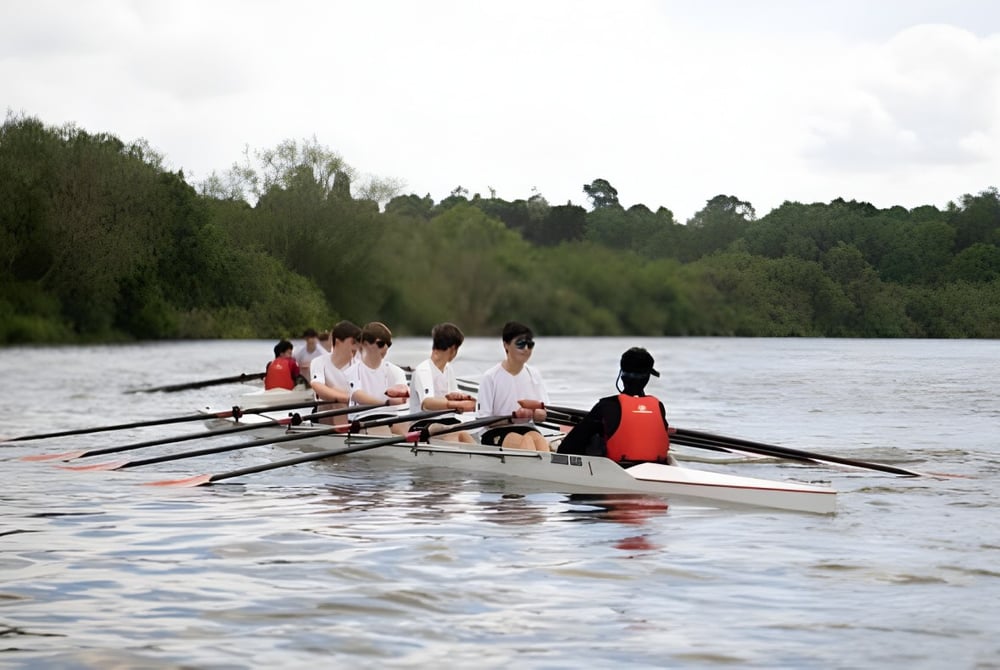 Eine Gruppe von Schülerinnen und Schülern des Cheltenham College rudert auf einem ruhigen Fluss, umgeben von bewaldeter Landschaft.