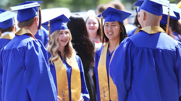 Eine Gruppe von Absolventen der Chilliwack Secondary School steht mit blauen Talaren und Hüten gemeinsam vor grüner Kulisse.