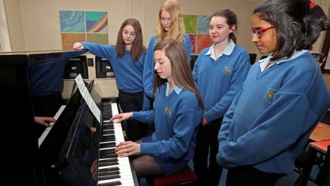 Eine Gruppe Schülerinnen und Schüler der Christ King Secondary School versammelt sich um ein Klavier in einem Raum mit Kunstwerken im Hintergrund.