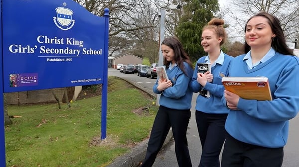 Drei Schülerinnen stehen vor einem Schild der Christ King Secondary School mit Bäumen und Wiese im Hintergrund.