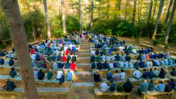 Eine große Gruppe von Menschen sitzt auf Holzbänken im Wald an der Christ School.