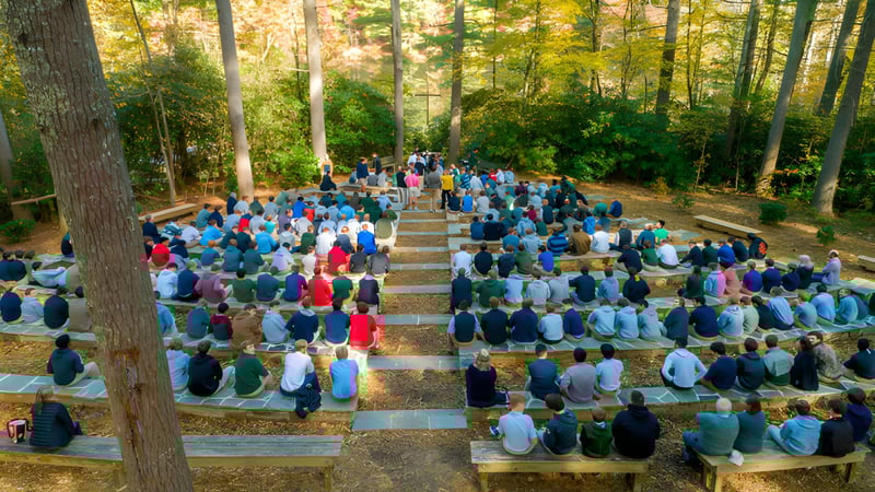 Eine große Gruppe von Menschen sitzt auf Holzbänken im Wald an der Christ School.