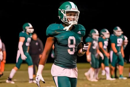 Ein Footballspieler der Christ School steht in einem grünen Trikot auf dem Spielfeld mit seinen Mannschaftskameraden im Hintergrund.