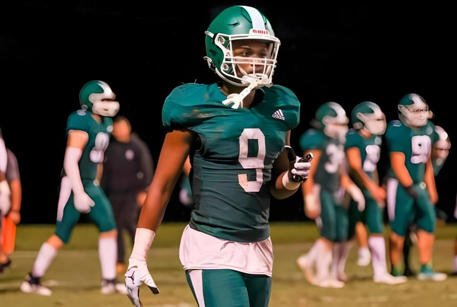 Ein Footballspieler der Christ School steht in einem grünen Trikot auf dem Spielfeld mit seinen Mannschaftskameraden im Hintergrund.