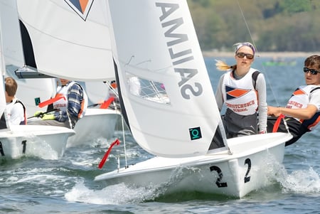 Zwei Schülerinnen und Schüler der Christchurch School segeln ein kleines Boot auf einem See vor bewaldetem Hintergrund.