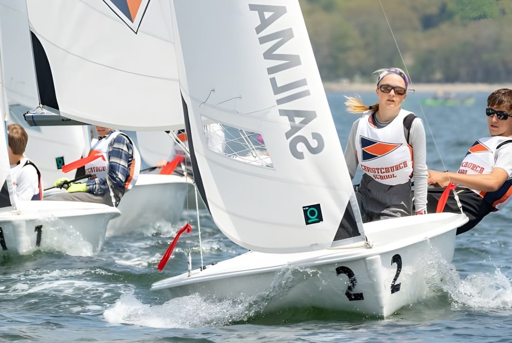 Zwei Schülerinnen und Schüler der Christchurch School segeln ein kleines Boot auf einem See vor bewaldetem Hintergrund.