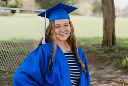 Eine Absolventin der Christian Life School steht im blauen Abschlussgewand lächelnd auf einer Wiese mit Bäumen im Hintergrund.