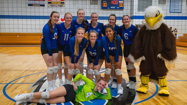 Eine Gruppe junger Volleyballspielerinnen in blauen Trikots posiert auf dem Basketballfeld der Christian Life School mit dem Maskottchen in einem gelben Kostüm.
