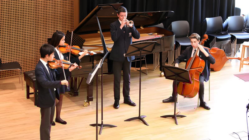 Schüler der Claremont School treten in formaler Kleidung mit verschiedenen Musikinstrumenten auf einer Bühne vor einer Holzpaneelwand auf.