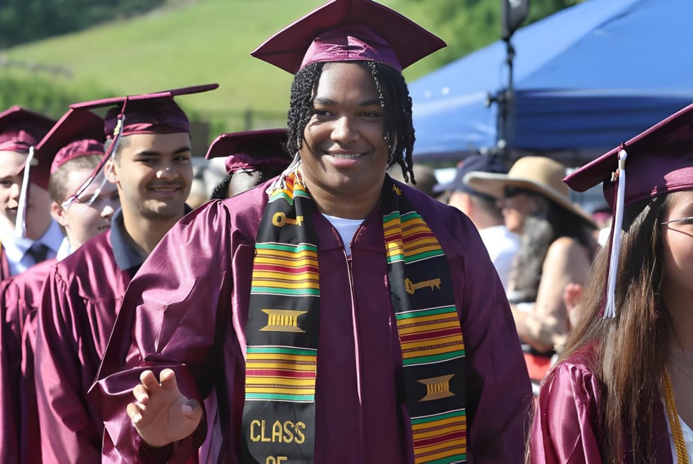 Absolventen in violetten Talaren der Claremont Secondary School stehen vor einem blauen Zelt und Bäumen.