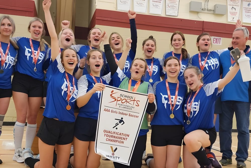 Schülerinnen der Clarenville High School feiern ihren Sieg mit Medaillen und Pokal vor einer rot gestrichenen Wand.