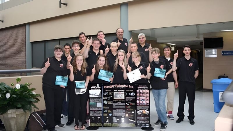 Eine Gruppe Schüler steht vor dem Gebäude der Clarenville High School und hält verschiedene Gegenstände sowie ein Schild.