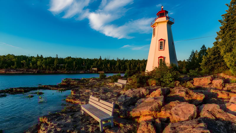 Ein Leuchtturm steht auf einem felsigen Vorsprung vor einem ruhigen See mit Wald im Hintergrund auf dem Gelände der Clarke Road Secondary School.