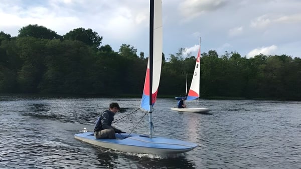 Eine Person segelt auf einem kleinen Segelboot auf dem Wasser beim Clifton College vor bewölktem Himmel und Bäumen.