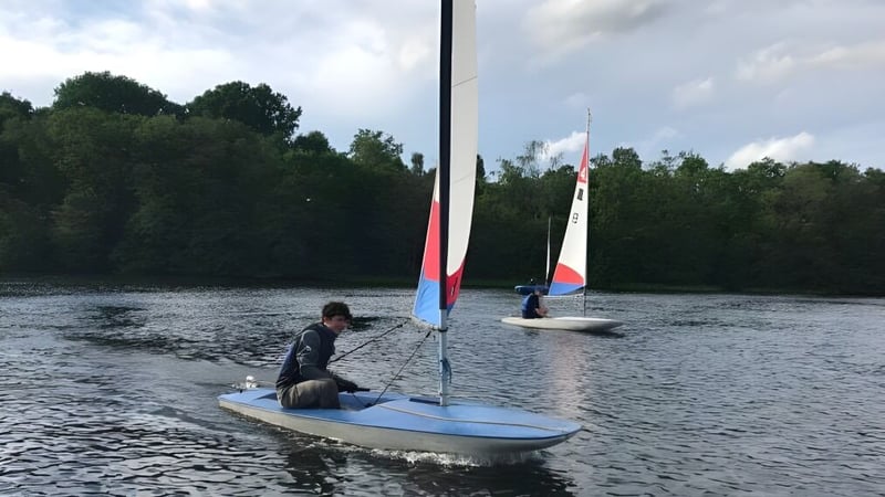 Eine Person segelt auf einem kleinen Segelboot auf dem Wasser beim Clifton College vor bewölktem Himmel und Bäumen.
