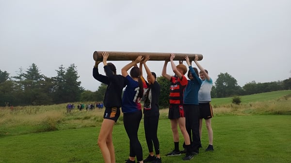 Schüler des Clonfert College heben gemeinsam einen großen Baumstamm auf einer Wiese mit Bäumen.