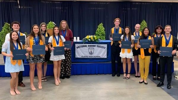 Eine Gruppe junger Frauen in Abschlusskleidung steht vor einem Banner der National Honor Society auf dem Campus der Clovis High School und hält ihre Diplome.