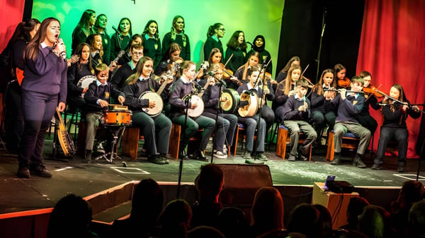 Der Chor der Cnoc Mhuire Secondary School tritt auf einer Bühne mit farbiger Beleuchtung und rotem Vorhang auf.