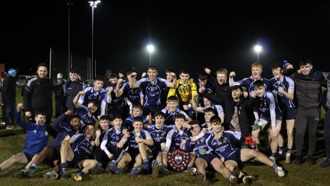 Eine Gruppe von Schülerinnen und Schülern der Cnoc Mhuire Secondary School steht bei Nacht auf dem Sportfeld unter hellen Flutlichtern.