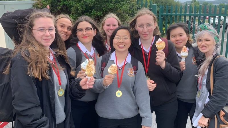 Schüler der Colaiste Muire (Cobh) in Abschlusskleidung versammeln sich mit Diplomen vor einem grünen Zaun.