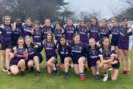 Eine Gruppe junger Fußballspielerinnen posiert auf dem Sportplatz der Colaiste Muire Mathair in violetten Trikots.
