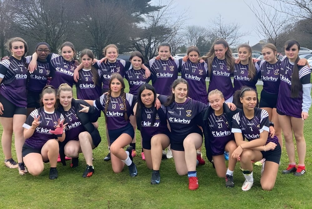 Eine Gruppe junger Fußballspielerinnen posiert auf dem Sportplatz der Colaiste Muire Mathair in violetten Trikots.