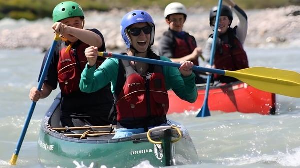 Schüler der Coláiste Bhaile Chláir paddeln mit bunten Helmen in einem kleinen grünen Boot auf einem Fluss durch eine grasbewachsene Landschaft.