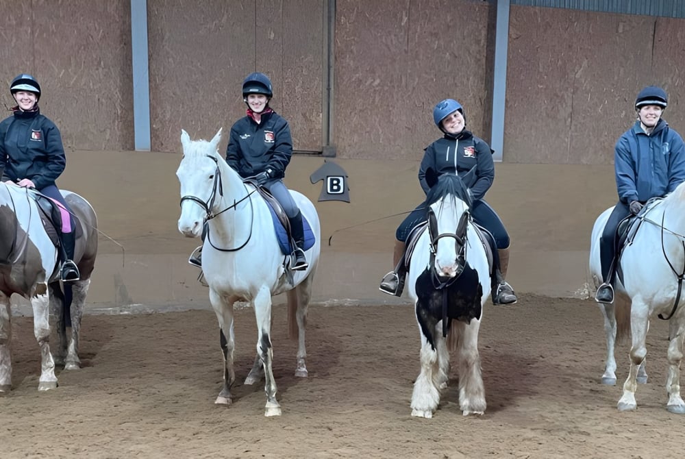 Schüler der Colaiste Chiarain Athlone reiten in einer Halle mit Boden aus brauner Erde und Holzwänden.