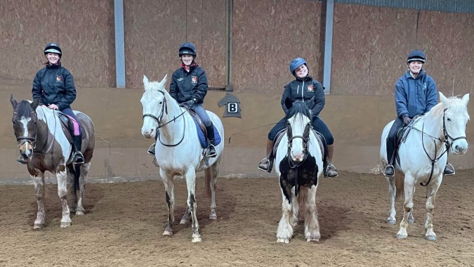 Vier Schüler der Colaiste Chiarain Athlone reiten in einer Indoor-Reithalle mit Holzwänden.